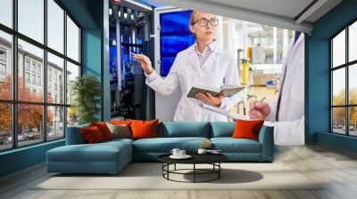 Portrait shot of attractive technician wearing white coat and eyeglasses trying to adjust equipment at production department of milk plant with help of male colleague Wall mural