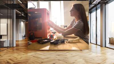 Profile view of beautiful young engineer wearing striped T-shirt sitting at office desk and using 3D printer, lens flare Wall mural