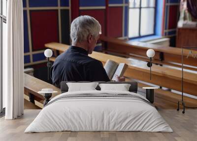 Rear view of senior priest reading Bible during praying while sitting on bench in church Wall mural