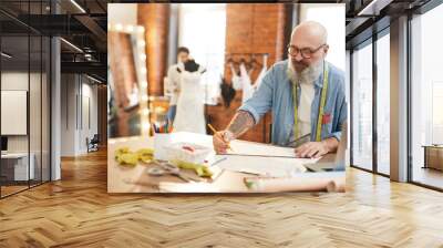 Senior tailor or fashion designer making sketch of new items for seasonal collection by workplace Wall mural