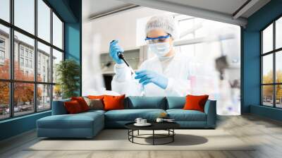 Serious concentrated female microbiologist in sterile clothing and safety goggles sitting at table and dropping reagent in petri dish while doing research in laboratory Wall mural