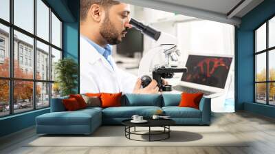 Serious concentrated young Arabian microbiologist in white coat sitting at table with laptop and flask and studying organism using microscope Wall mural