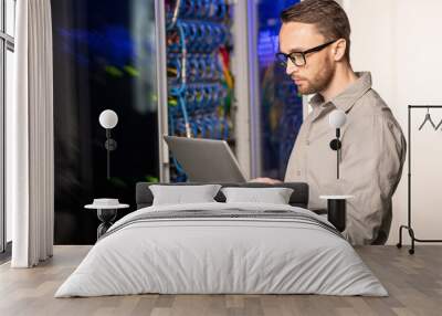 Serious thoughtful handsome young server engineer in glasses standing in database center and using laptop while troubleshooting problem with network Wall mural