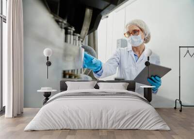 Shot from pill press machine of female process technician in sterile classroom clothing holding digital tablet while inspecting manufacturing equipment at pharmaceutical factory, copy space Wall mural