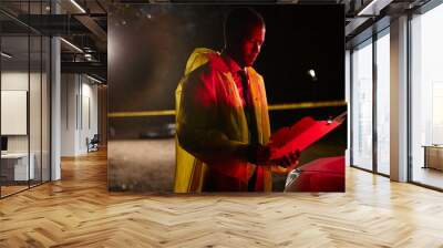 Side view portrait of African American police office holding folder and reading documents outdoors at crime scene with red siren lights, copy space Wall mural