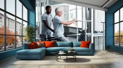 Side view portrait of senior man doing rehabilitation exercises in gym at medical clinic with therapist assisting Wall mural