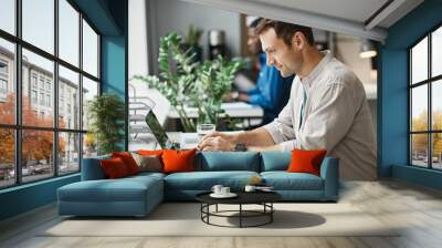 Side view portrait of smiling adult man using laptop while enjoying work in office, copy space Wall mural