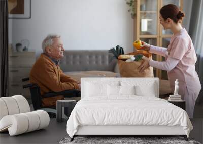 Side view portrait of smiling senior man in wheelchair looking at female nurse bringing groceries, assistance and food delivery concept Wall mural