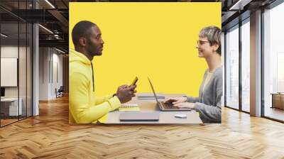 Side view portrait of two contemporary business people sitting opposite each other at desk during meting and smiling against yellow background Wall mural