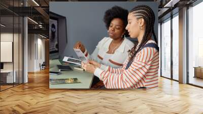 Side view portrait of two creative African American women discussing photos in studio and holding art prints for selection Wall mural