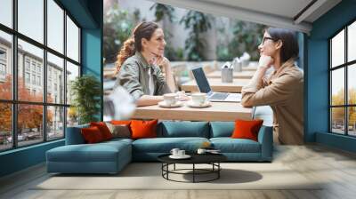 Side view portrait of two modern young women sitting at table in cafe while discussing business during meeting on outdoor patio, copy space Wall mural