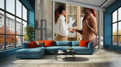 Side view portrait of two successful young women shaking hands and smiling cheerfully while standing in modern office interior, copy space Wall mural