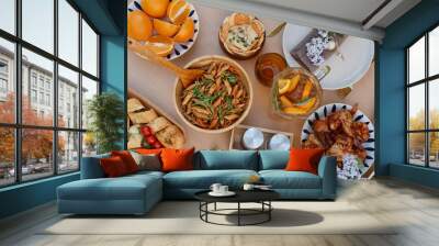 Top-down view of an elegant dining setup with Mediterranean dishes including pasta, chicken, assorted bread, fresh fruits, and beverage, arranged on a wooden table with flowers adding charm Wall mural