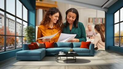 Two attractive stylish women choosing best design sketch, their colleagues coworking behind them Wall mural