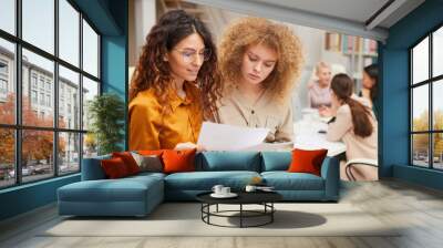 Two young businesswomen looking at documents, their colleagues coworking behind them Wall mural