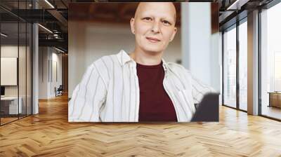 Vertical portrait of bald adult woman smiling at camera while holding laptop and working at home in modern interior, alopecia and cancer awareness Wall mural