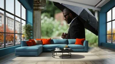 Vertical shot of Black man grieving over loss of relative at cemetery next to grave holding white rose to commemorate deceased and hiding from rain under black umbrella, copy space Wall mural