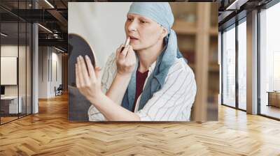 Vertical warm-toned portrait of confident bald woman putting on makeup and lipstick while looking in mirror at home, embracing beauty, alopecia and cancer awareness Wall mural