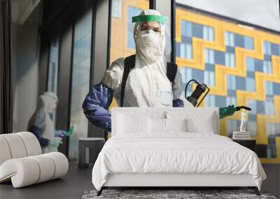 Waist up portrait of female worker wearing hazmat suit and holding disinfecting equipment looking at camera while standing in office, copy space Wall mural