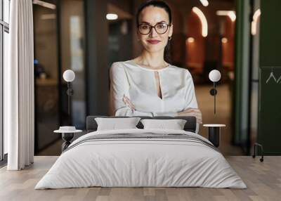 Waist up portrait of smiling Caucasian brunette businesswoman in glasses crossing arms and looking at camera standing in office building Wall mural