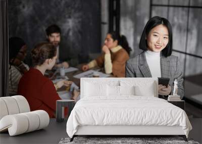 Waist up portrait of successful Asian businesswoman smiling at camera with diverse group of people meeting in background, copy space Wall mural