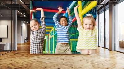 Waist up portrait of three happy kids jumping raising hands while having fun in play area and looking at camera Wall mural