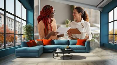 Waist up portrait of two successful young businesswomen discussing documents and smiling cheerfully while standing in modern office interior, copy space Wall mural