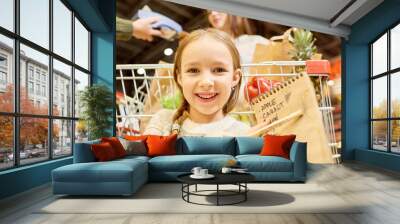 Warm-toned portrait of happy little girl grocery shopping in supermarket, smiling looking at camera and pointing at shopping list with food items Wall mural