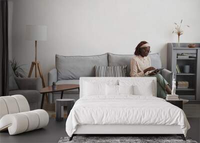 Wide angle portrait of young African-American woman using digital tablet while sitting on sofa at home in minimal grey interior, copy space Wall mural