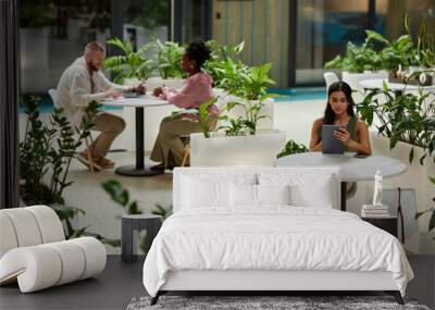 Wide angle shot of woman using tablet while sitting at table in cafe of office building. Business people working in cozy cafe featuring modern biophilic design Wall mural