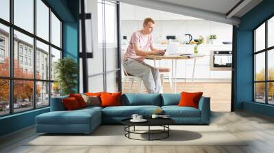 Young casual man sitting by table in the kitchen in front of laptop while networking Wall mural