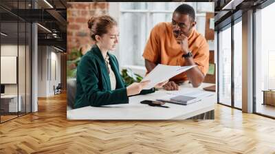 Young confident female economist or lawyer checking documentation by desk and consulting African-American male colleague Wall mural