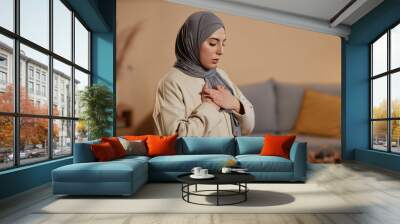 Young Islamic woman in grey hijab and beige sweatshirt expressing gratitude to her God during pray while keeping her hands on chest Wall mural