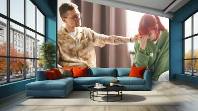 Young man with tattoos comforts young woman who is visibly upset, sitting indoors. Providing emotional support while woman covers face with hand, showing deep emotions Wall mural