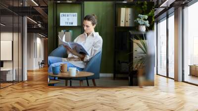 Young manager or economist in casualwear sitting in comfortable armchair and looking through manual in openspace office Wall mural