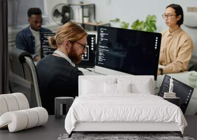 Young serious diversity programmer typing on laptop keyboard while sitting in front of computers by workplace against two coworkers Wall mural