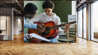 Youthful diligent girl playing acoustic guitar while sitting in front of African American male teacher consulting her at lesson Wall mural