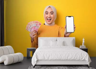 Beautiful amazed Asian girl wearing hijab showing mobile phone with blank white screen, holding cash money isolated on yellow background, mockup, copy space Wall mural