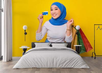 Cheerful young Asian Muslim woman holding credit card and shopping bags, looking at camera isolated on yellow background Wall mural