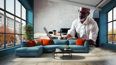 Adult stylish businessman wearing a classic glasses and working at the wood table in modern loft studio.Bearded middle age man in white shirt making notes notebook. Horizontal,blurred background. Wall mural