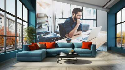 Bearded pensive coworker working at sunny loft office on laptop while sitting at wooden table.Businessman reading documents on notebook display.Blurred background,horizontal. Wall mural