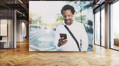 Happy american African man using smartphone outdoor.Portrait of young black cheerful man texting a sms message while listining to music.Blurred background, horizontal.Flare effects. Wall mural