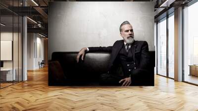 Portrait of adult businessman wearing trendy suit and sitting modern studio on leather sofa against the empty concrete wall. Horizontal, space for ad Wall mural