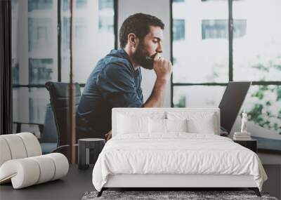 Young pensive coworker working at sunny work place loft while sitting at the wooden table.Man analyze document on laptop display.Blurred background.Horizontal. Wall mural