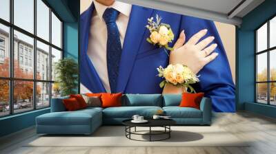 The hand of the bride with a boutonniere rests on the forearm of the groom in a blue suit with a boutonniere and tie Wall mural