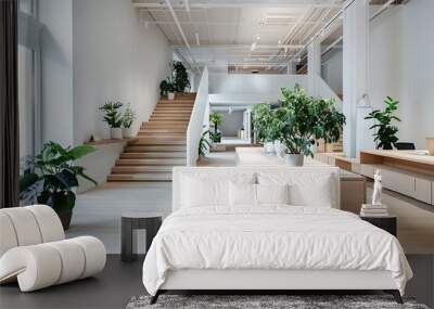 Bright, minimalist office with wooden desks, light gray walls, and a white ceiling. Green desk plants add vibrancy, while a staircase to the second floor completes the space. Wall mural