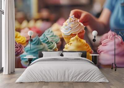 A closeup of a table full of colorful and decorated cupcakes. A baker is in the background, putting the finishing touches on the last cupcake. Wall mural