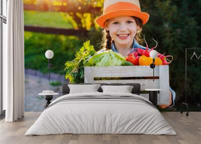 Kids little girl holding a basket of fresh organic vegetables in the background of a home garden at sunset. Healthy family lifestyle. Harvest time in autumn. The child the farmer. Wall mural