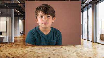 Handsome serious school age boy in teal t shirt looking at camera isolated on brown background Wall mural