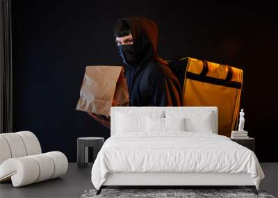 delivery man in black clothes in medicine mask with a yellow backpack and paper bag on a black background with orange light Wall mural